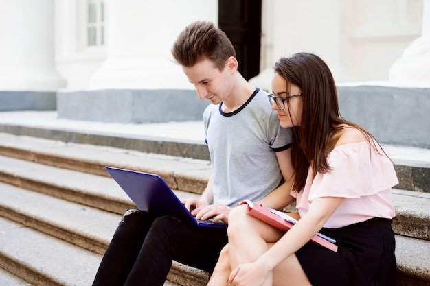 Due allegri studenti adolescenti che fanno i compiti con il quaderno, la cartella e il laptop si siedono all'aperto sulle scale dell'università