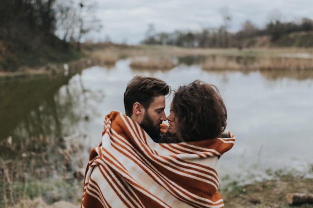 Due amanti caucasici avvolti in una coperta in riva al lago. La giovane coppia sta abbracciando il giorno di autunno all'aperto. San Valentino. Concetto di amore e famiglia.