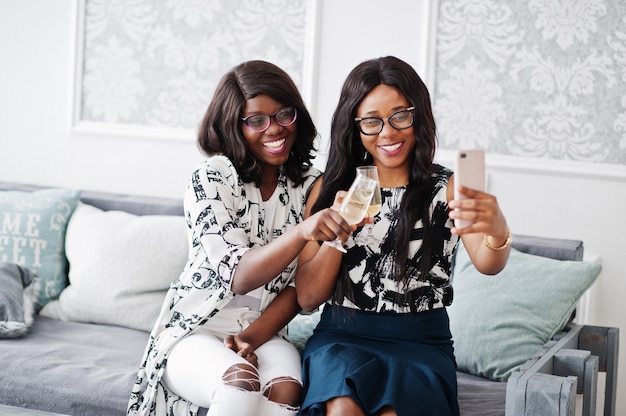 Due amiche africane indossano occhiali posati nella stanza bianca interna, bevendo champagne e facendo selfie.