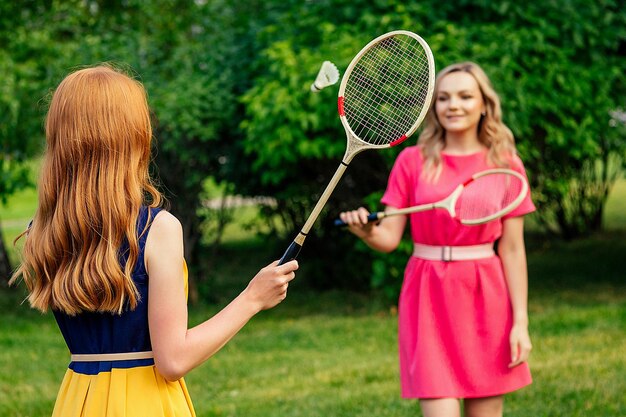 Due amiche allegre divertenti bella giovane donna irlandese di zenzero rossa in un vestito giallo e persona di sesso femminile bionda europea in un vestito rosa giocando a tennis con racchetta da badminton nel parco estivo