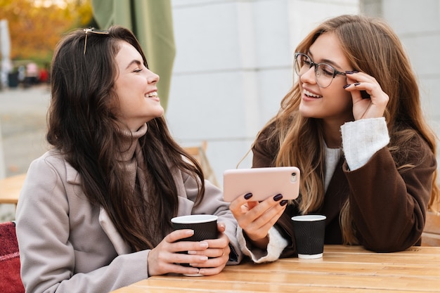 Due amiche allegre e attraenti sedute al bar all'aperto, guardando il cellulare mentre bevono caffè