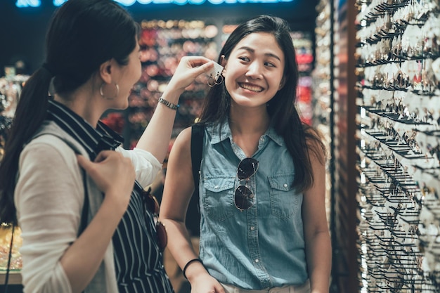 due amiche asiatiche piuttosto giovani che provano un paio di orecchini mentre fanno acquisti insieme in gioielleria. ragazza che aiuta la sorella sorridente a scegliere gli accessori nel negozio di moda. le donne felici trascorrono il tempo libero