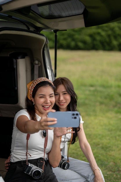 Due amiche asiatiche spensierate sono sedute nel bagagliaio aperto dell'auto e usano il telefono per farsi dei selfie