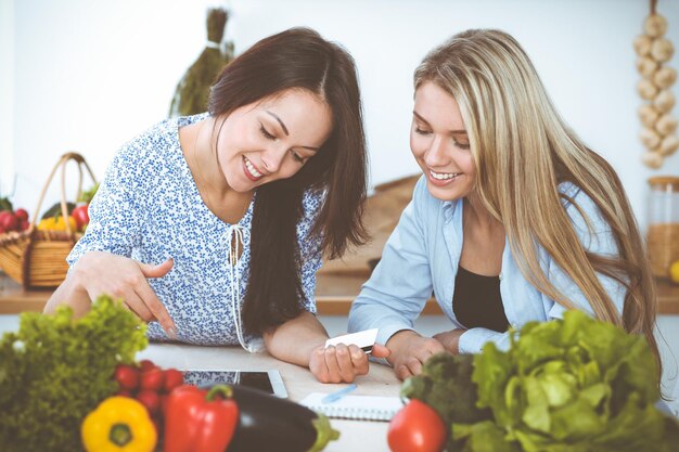 Due amiche che scelgono la ricetta per un pasto delizioso seduti al tavolo in cucina Tablet pc è il miglior libro di cucina