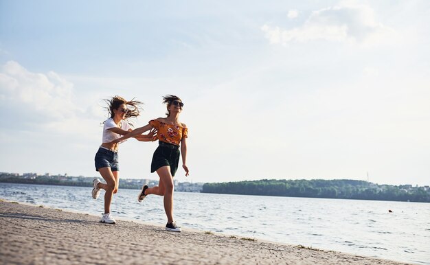 Due amiche corrono e si divertono in spiaggia vicino al lago durante il giorno soleggiato.