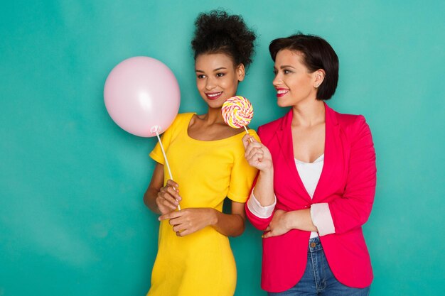 Due amiche in abiti colorati a sfondo blu. Donne afro-americane e caucasiche che si divertono, tengono in mano un palloncino e un lecca-lecca, offrendo caramelle ad un amico. Condivisione, concetto di amicizia multietnica