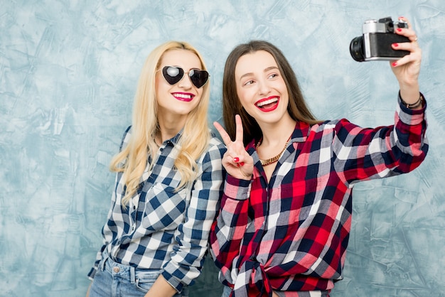 Due amiche in camicie a scacchi e jeans che fotografano con una fotocamera retrò sullo sfondo del muro dipinto di blu