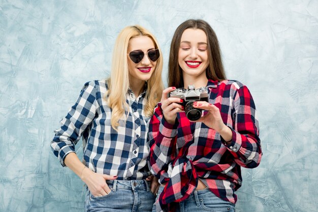 Due amiche in camicie a scacchi e jeans che fotografano con una fotocamera retrò sullo sfondo del muro dipinto di blu