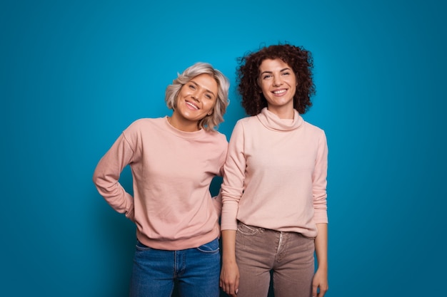 Due amiche in posa su sfondo blu una ragazza con i capelli biondi corti e lisci ma l'altra con...