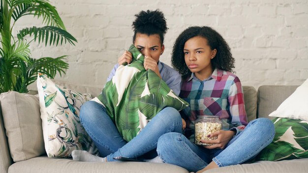 Due amiche ricce di razza mista sedute sul divano e guardano un film molto spaventoso in TV a casa