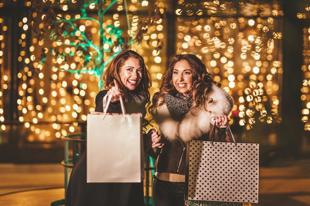 Due amiche si godono la notte facendo shopping e ridendo con le borse della spesa in mano.