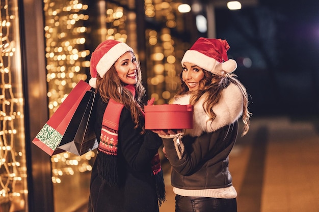 Due amiche si godono la notte facendo shopping, si guardano e ridono con le borse della spesa e la scatola rossa in mano.