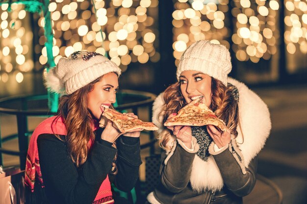 Due amiche si godono la serata fuori, guardandosi e ridendo con fette di pizza in mano.