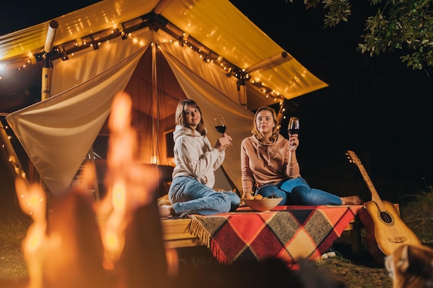 Due amiche sorridenti che bevono vino e mangiano frutta seduti in un'accogliente tenda glamping in falò serale autunnale Tenda da campeggio di lusso per vacanze all'aperto e vacanze Concetto di stile di vita