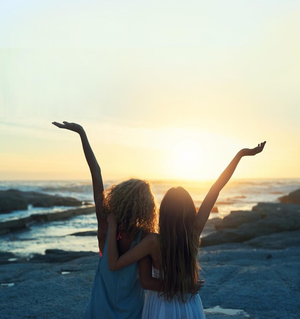 Due amiche sulla spiaggia che celebrano con le braccia alzate guardando il tramonto godendosi lo stile di vita della libertà di viaggio