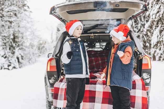 Due amici adolescenti ragazzi con cappelli rossi da Babbo Natale con scintillii fuoco del Bengala in piedi vicino al bagagliaio dell'auto decorato per Natale e Capodanno nella foresta invernale innevata Viaggio su strada e viaggi locali