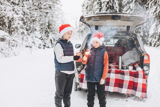 Due amici adolescenti ragazzi con cappelli rossi da Babbo Natale con scintillii fuoco del Bengala in piedi vicino al bagagliaio dell'auto decorato per Natale e Capodanno nella foresta invernale innevata Viaggio su strada e viaggi locali