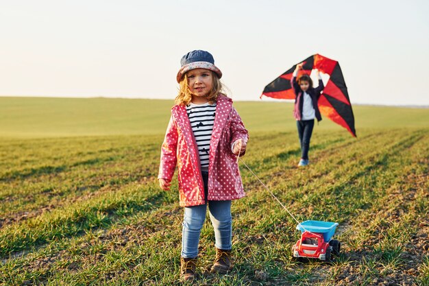 Due amici di bambine si divertono insieme con l'aquilone e l'automobile del giocattolo sul campo al giorno soleggiato