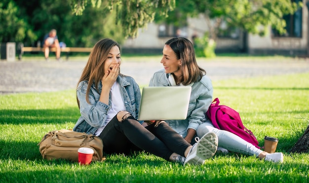 Due amici di ragazza felice bella giovane studentessa in abiti casual in denim sono rilassanti nel parco del college con laptop e smartphone dall'università e bere caffè