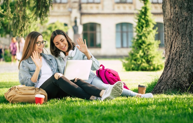 Due amici di ragazza felice bella giovane studentessa in abiti casual in denim sono rilassanti nel parco del college con laptop e smartphone dall'università e bere caffè