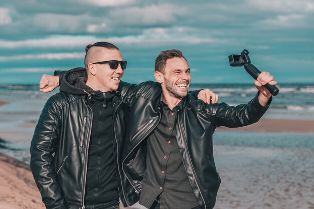 Due amici sorridenti belli che fanno Selfie utilizzando la macchina fotografica di azione con stabilizzatore cardanico in spiaggia.