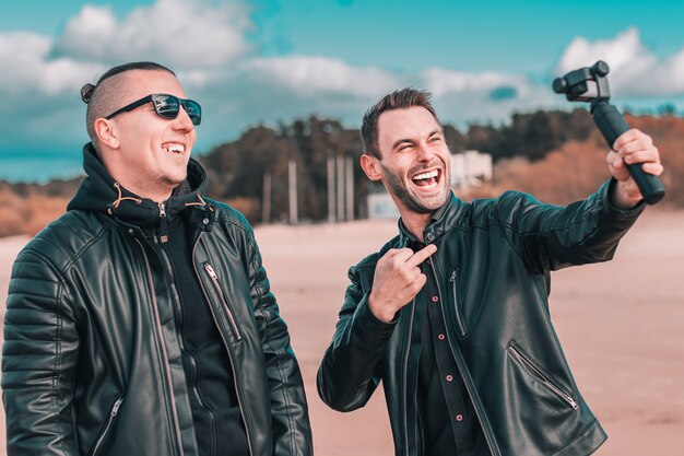 Due amici sorridenti belli che fanno selfie utilizzando la macchina fotografica di azione con stabilizzatore cardanico in spiaggia