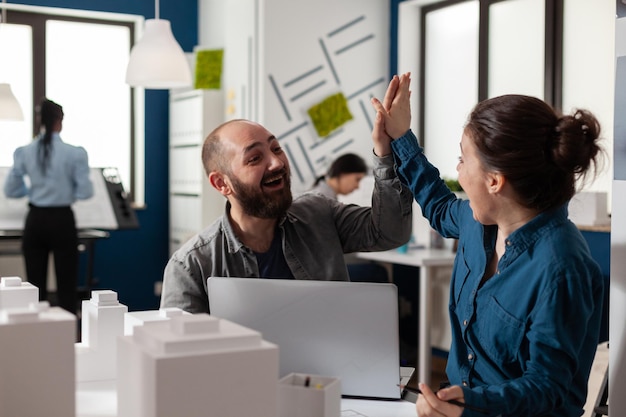 Due architetti in alto cinque seduti alla scrivania in un ufficio architettonico moderno con laptop e modello in scala di schiuma di edifici residenziali. Colleghi sorridenti felici di successo che celebrano il progetto di squadra.