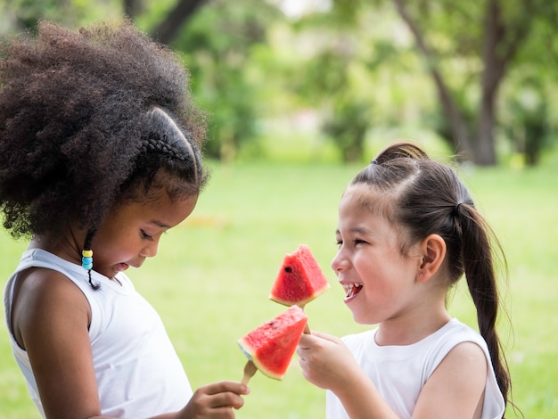 Due bambine adorabili di varie etnie che si divertono a mangiare anguria nel parco.