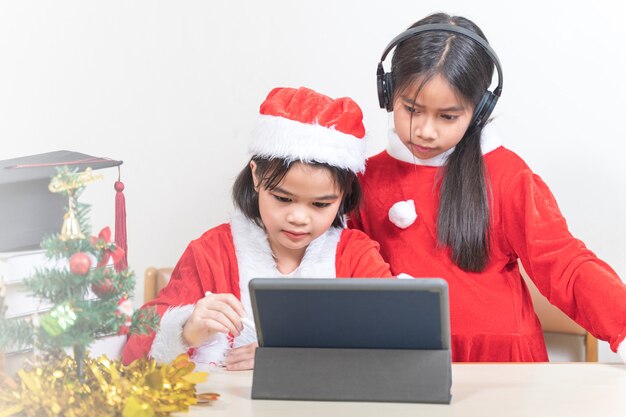 Due bambine asiatiche indossano Babbo Natale chiacchierando con un amico su tablet digitale. Concetto di vacanza di Natale Stock Photo