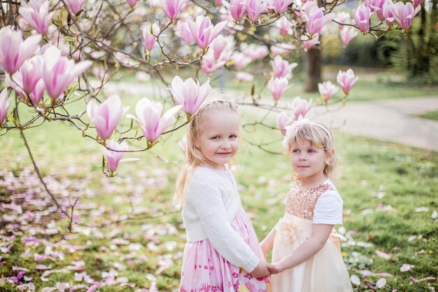 Due bambine bionde carine di 3 anni stanno giocando nel parco vicino a una magnolia in fiore. Bere il tè. Pasqua. Primavera.