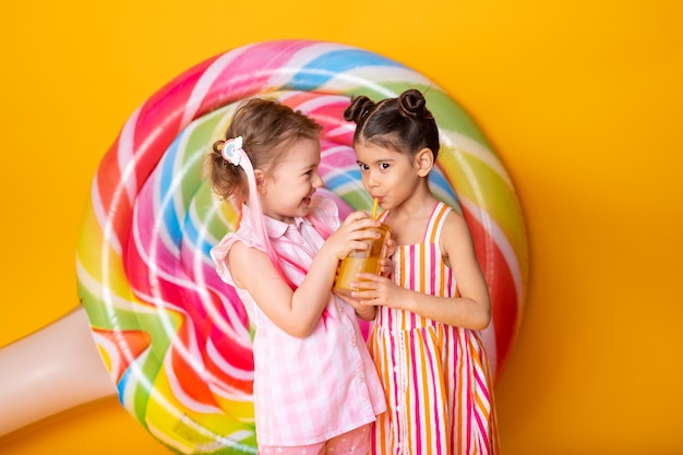 Due bambine felici in vestito colorato che bevono succo d'arancia divertendosi sulla superficie gialla con lecca-lecca.