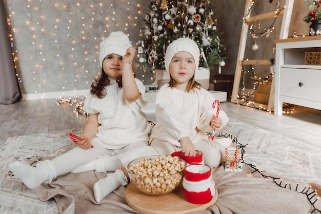 Due bambine in cappelli lavorati a maglia invernali sono sedute sul pavimento vicino all'albero di Natale e mangiano popcorn. concetto di natale.