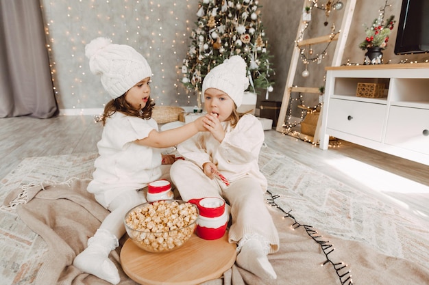 Due bambine in cappelli lavorati a maglia invernali sono sedute sul pavimento vicino all'albero di Natale e mangiano popcorn. concetto di natale.