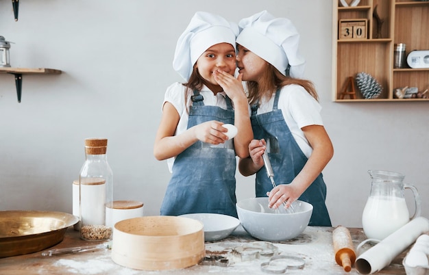 Due bambine in uniforme blu da chef che si parlano di segreti quando preparano il cibo in cucina