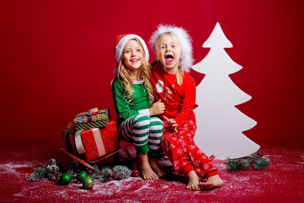 Due bambine sui cappelli di Natale su uno sfondo rosso si siedono vicino a un albero di Natale bianco con doni