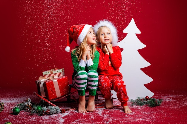 Due bambine sui cappelli di Natale su uno sfondo rosso si siedono vicino a un albero di Natale bianco con doni