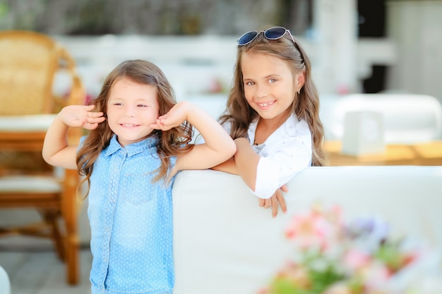 Due bambine sveglie che stanno in saltatori e vetri di sole sui precedenti del terrazzo nello studio. Concetto di estate, divertimento, famiglia e vacanze. Due sorelle di moda in posa.