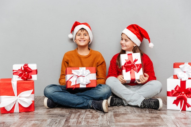 Due bambini allegri in cappelli di babbo natale seduti e in possesso di scatole regalo