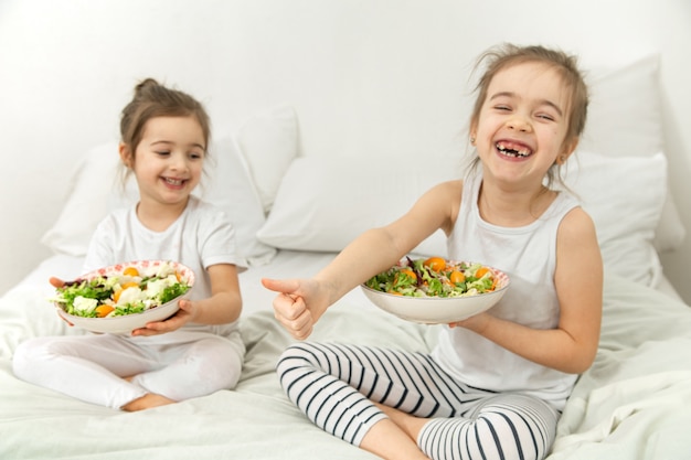 Due bambini carini felici che mangiano insalata di verdure nella camera da letto sul letto. Cibo sano per bambini e adolescenti.