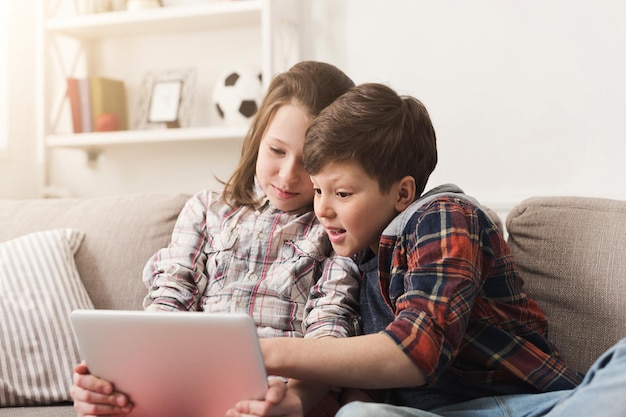 Due bambini con gadget. Sorella e fratello che condividono contenuti divertenti su tablet digitale sul divano di casa. Amicizia familiare e concetto di comunicazione