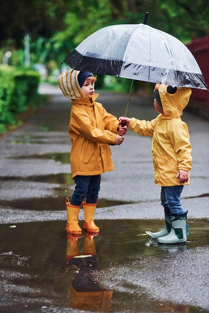 Due bambini con ombrello in mantelli impermeabili gialli e stivali che giocano insieme all'aperto dopo la pioggia