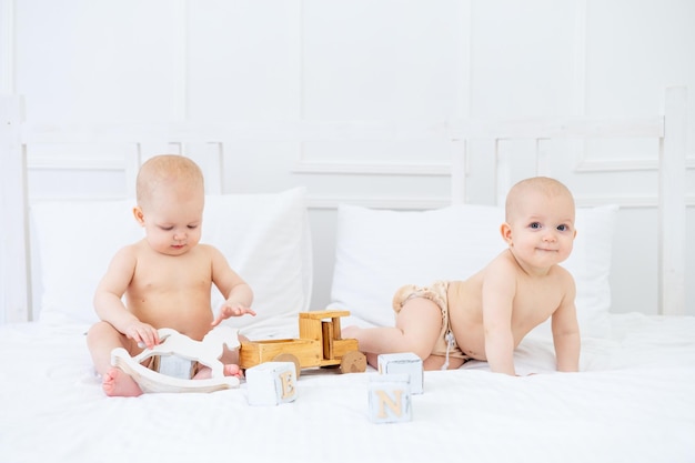 Due bambini gemelli di sei mesi in pannolini su un letto di cotone bianco in un body su un letto a casa giocando con giocattoli di legno e sorridendo