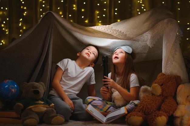 Due bambini piccoli giocano a casa la sera per costruire una tenda da campeggio dove leggere libri con una torcia e dormire all'interno. Concetto di: gioco, magia, creatività, sistemi di allarme