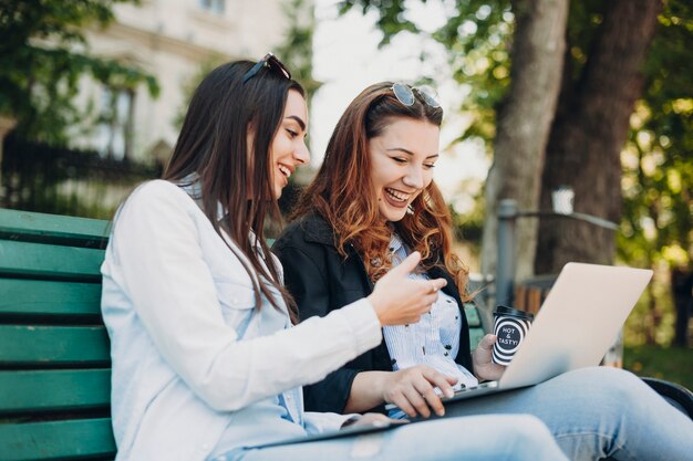 Due bei giovani amici che ridono mentre guardano lo schermo di un laptop seduto su una panchina nel parco a bere caffè.