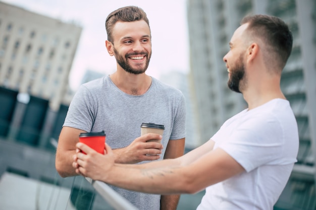 Due bei giovani migliori amici barbuti in abbigliamento casual sono in piedi all'aperto con tazze di caffè che ridono e hanno una conversazione.