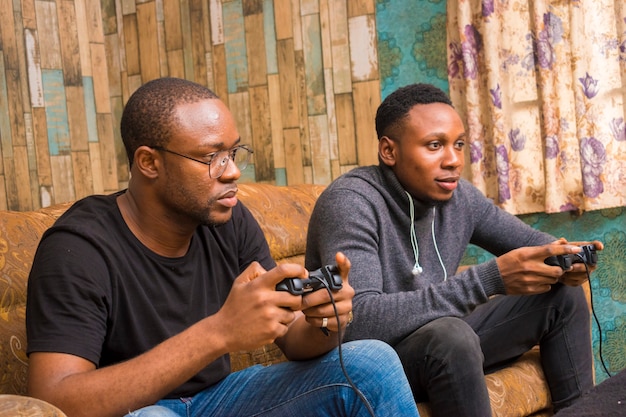 Due bei ragazzi africani seduti sul divano a giocare ai videogiochi con joystick, game pad, pad
