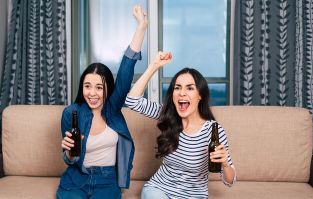 Due belle amiche allegre in abiti casual che guardano il calcio in TV seduti sul divano di casa a bere birre