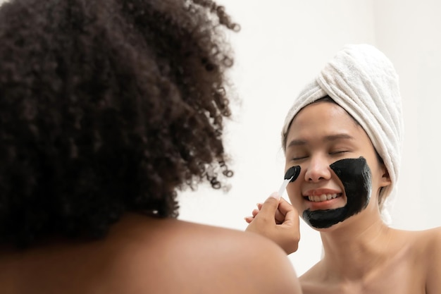 Due belle amiche in asciugamano che applicano maschera nutriente nera sul viso in camera da letto a casa trattamento viso cura della pelle trattamenti di bellezza cosmetici spa corpo sano maschera facciale concetto