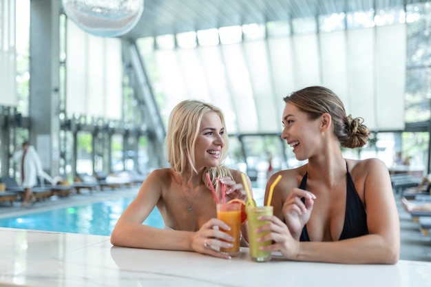 Due belle donne abbronzate sexy felici e alla moda alla moda con un cocktail, accanto alla piscina del resort