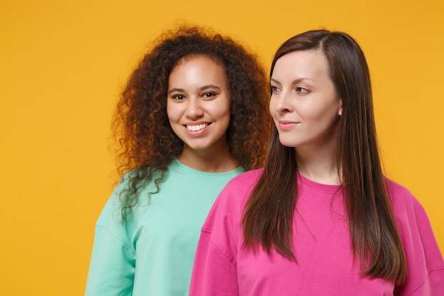 Due belle donne amiche europee e afroamericane ragazze in abiti rosa verdi in posa isolato su sfondo giallo arancio ritratto in studio. Concetto di stile di vita delle persone. Mock up spazio di copia.